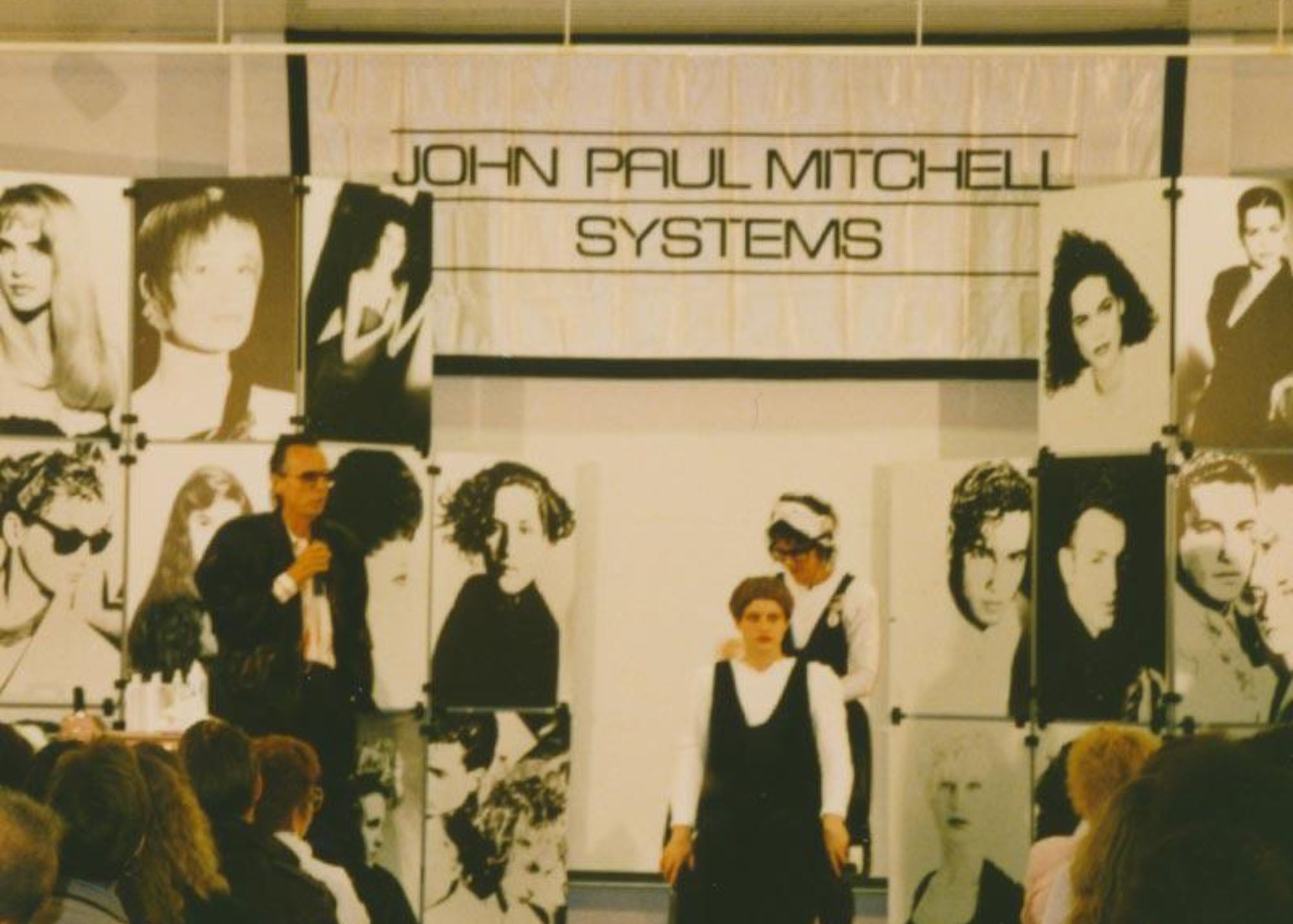 John Paul Mitchell Systems banner with black and white headshots of people while Paul Mitchell speaking on stage.