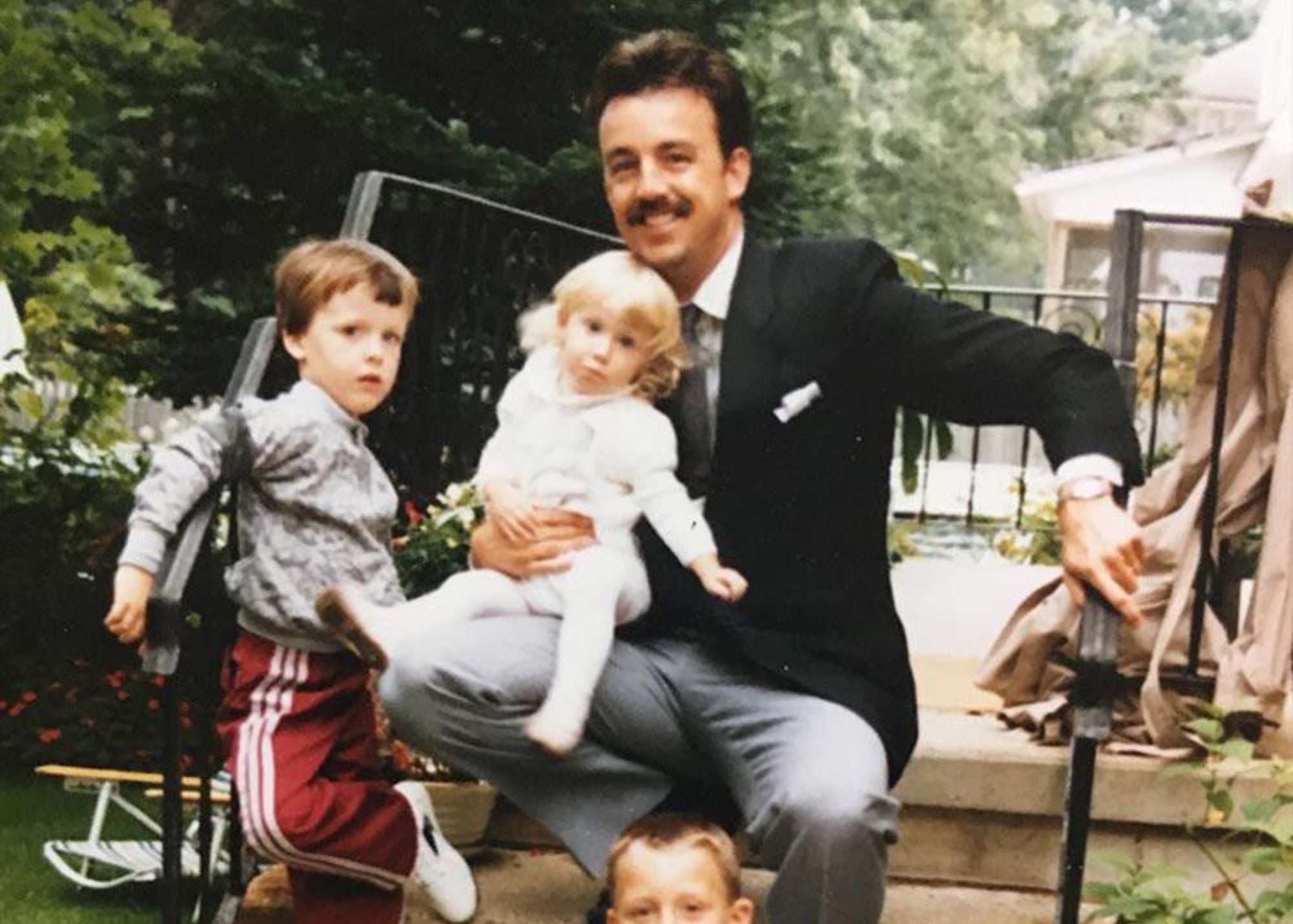 Man in a suit holding two children outdoors with a garden background