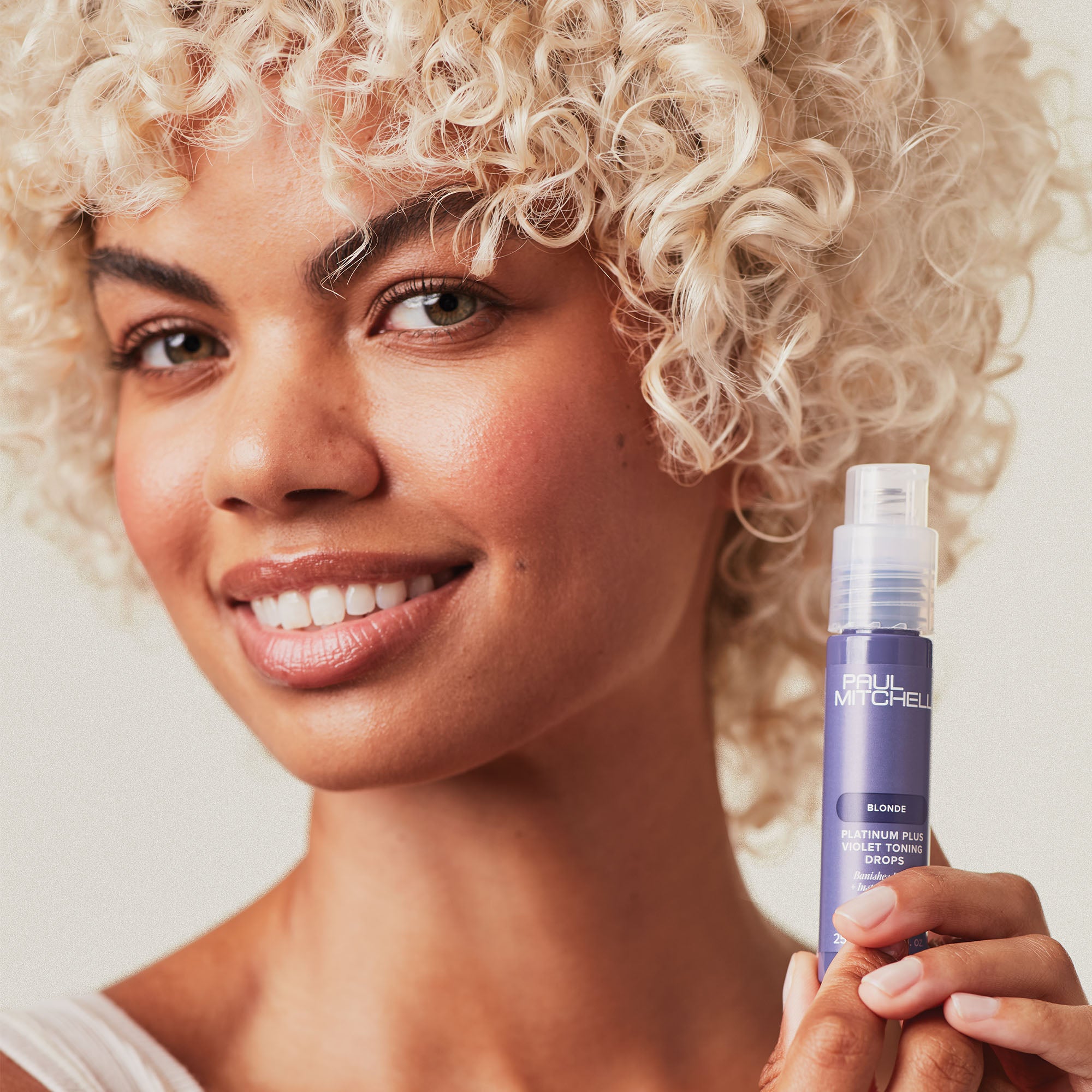 Image of Paul Mitchell Blonde female model with blonde curly hair holding bottle of toning drop near her face
