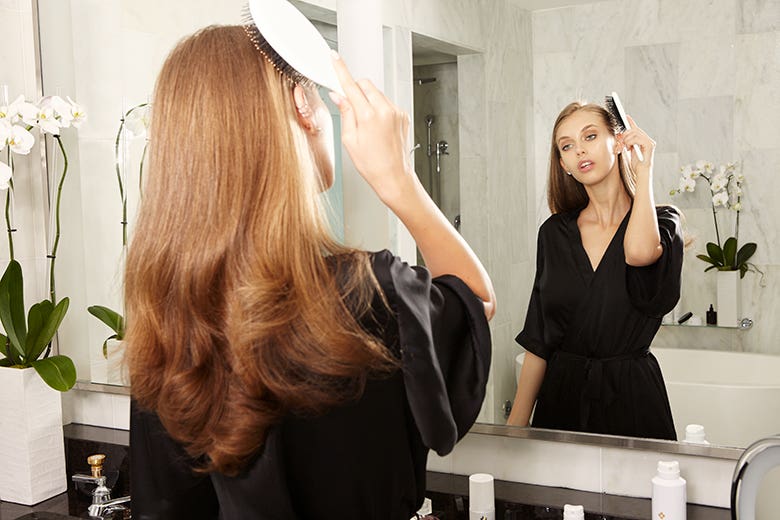Marula How-To: Glamorous Waves Step 1 of 11. Woman standing in front of mirror brushing her hair.