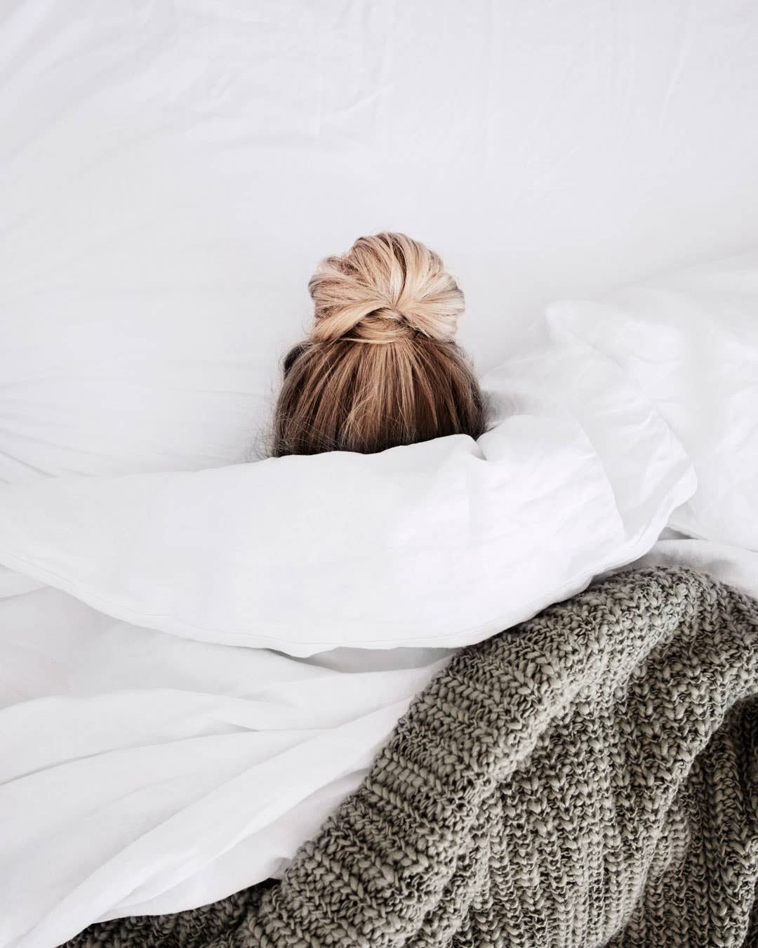 Model in bed with the covers pulled up, only showing her head with a blonde bun sticking out.